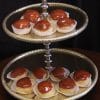 A two-tiered silver tray displays Caramel Cream Puffs, each in a white paper cup and topped with glossy caramel glaze. The dark background highlights the delicate treats.