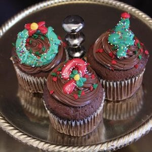 Three CH-064 Display Christmas Decor Chocolate cupcakes with white filling are topped with festive green and red holiday designs—a wreath, a tree, and a candy cane—and arranged on a silver cake stand.