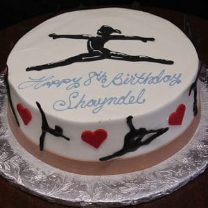 The CB-026B is a round birthday cake with white icing, black gymnast silhouettes, red hearts, blue "Happy 8th Birthday Shayndel" script, and a gymnast in a split leap on top.