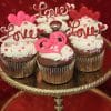 The VC-036 Display Valentine chocolate cupcakes with white filling feature pink and red heart sprinkles and “Love” toppers, beautifully arranged on a gold cake stand against a red background.