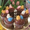 EA-040 Display Easter Decor chocolate cupcake with white filling features colorful Easter-themed toppers like candy eggs, ducks, and bunnies, arranged on a gold cake stand with pink tulips and flowers in the background.