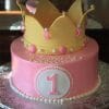 The CB-016D cake features pink icing, a large yellow crown topper, white and pink pearls, a white circle with the number 1 on the front, and "Victoria" written in white icing.