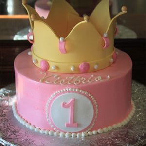 The CB-016D cake features pink icing, a large yellow crown topper, white and pink pearls, a white circle with the number 1 on the front, and "Victoria" written in white icing.