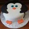 The CB-012A is a round cake crafted to resemble a cartoon penguin, featuring black wings, big blue eyes, an orange beak and feet, presented on a silver patterned cake board.