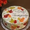 The THX-024 is a round cake decorated with autumn leaves and green swirls, topped with "Happy Thanksgiving" in cursive icing, and set on a foil-covered board. Orange flowers are seen in the background.