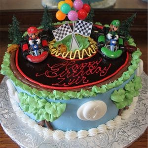 The SC-008 Mario cake is round and Mario Kart-themed, with Mario and Luigi in go-karts, checkered flags, balloons, green trees on top, and "Happy Birthday Ziyir" written in red icing.