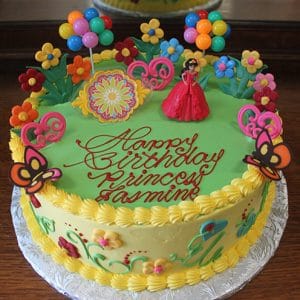 A round birthday cake named SC-019 Elena, decorated with colorful flowers, balloons, butterflies, and a princess figurine. "Happy Birthday Princess Jasmine" is written in red icing on top.