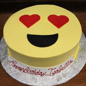 The CB-005C is a round cake featuring a yellow emoji face with heart-shaped eyes and a wide smile, placed on a silver board with a "Happy Birthday Isabella" banner in red icing.
