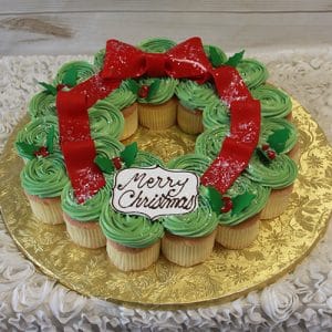 The CC-081 Christmas Wreath Gold features cupcakes with chocolate filling, decorated with green frosting, red bows, holly accents, and a "Merry Christmas" plaque, all displayed on a gold board.