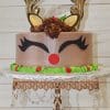 CH-023 is a round cake designed as a reindeer with antlers, ears, closed eyes with long lashes, a red nose, chocolate frosting hair, green icing at the base, and red berries on an ornate stand.