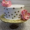 The BR-020 is a white cake with gold polka dots and two big pink sugar flowers, presented on a gold stand with crystal accents. It’s set against light wood and a white ruffled background.