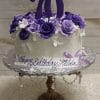 The BD-060H Shades of Purple Flowers cake is white, decorated with purple and white flowers, topped with a large purple 70, displayed on a stand, with a plaque reading "Happy Birthday Hilda" in script.