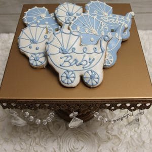 The CE-004c baby shower features sugar cookies shaped like carriages with blue and white icing, some labeled "Baby," displayed on a gold stand with crystal accents atop a white floral surface.