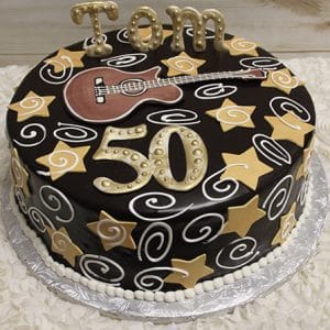 BD-063 Guitar/gold stars: Round chocolate cake with yellow stars, white swirls, a fondant guitar, and “Tom 50” in large gold letters on top, presented on a silver platter.