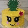 CB-020A is a round cake designed as a cute pineapple, featuring yellow frosting, a fondant green leaf topper, big cartoon eyes, pink cheeks, a smiling mouth, and a pink flower accent on the side.