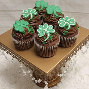 Six STP-008 St. Patrick's Day Chocolate cupcakes with chocolate Baileys filling are topped with green and white shamrock decorations and displayed on a gold stand with hanging crystal beads against a textured white background.