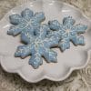 Three AP-075 snowflake cookies with light blue and white icing are arranged on a white, scalloped-edge plate placed on a textured, cream-colored surface.