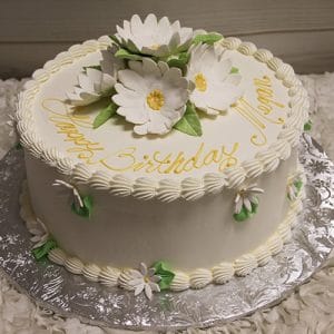 The BR-028 is a round white cake with white and yellow daisies, green leaves, piped borders, and "Happy Birthday Megan" written in yellow icing on top. It is presented on a silver foil-covered board.