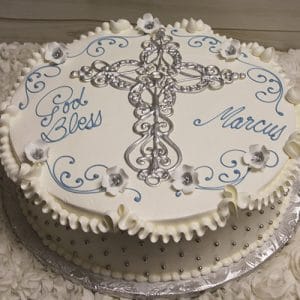 RC-023 is a round white cake with a silver cross, blue swirls, white flowers, "God Bless Marcus" inscription, ruffled edges, and silver edible pearls around the sides.