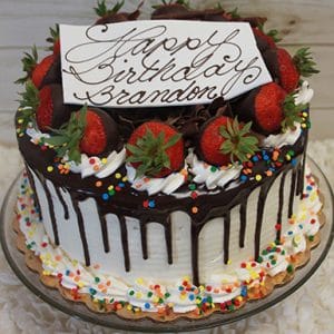 The BD-007 is a chocolate-dripped birthday cake with strawberries, whipped cream, and colorful sprinkles. A white plaque says “Happy Birthday Brandon” in cursive. The cake sits on a glass stand against a light wood background.