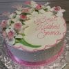 The BR-044 cake is round, adorned with white and pink flowers, pink piped dots, and a pink border. "Best Wishes Emma" is written in pink icing on top. The cake is presented on a silver floral-patterned board.