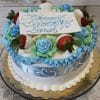 AN-000C is a round cake with white and blue icing, blue-green frosting, blue roses, chocolate-dipped strawberries, and a card that reads "Happy Birthday Sarah" in blue script. The cake is displayed on a glass stand.