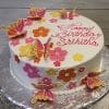 The CB-024B is a round white birthday cake adorned with pink, yellow, and orange flowers and butterflies, featuring “Happy Birthday Srihitha” in pink icing on top. It is presented on a silver tray.