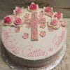 RC-052: A round white cake topped with pink and white frosting roses, a cross outlined in pink at the center, and “God Bless Caitlin & Julianne” written in pink cursive.