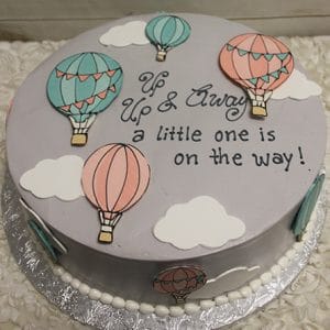 BB-065 is a round cake with pastel hot air balloons, white clouds, and the message “Up & Away a little one is on the way!” on light gray fondant—perfect for baby showers.
