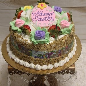 A carrot cake with “Happy Easter” on a pink plaque, topped with strawberries and colorful icing roses, edged with nuts, served on a gold cake board and ornate stand.