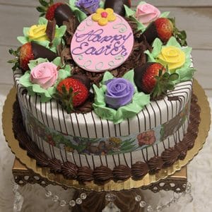The Strawberry Grand Marnier cake with Easter Decor features chocolate-dipped strawberries, pastel icing roses, green icing leaves, and a large pink oval reading "Happy Easter," all presented on a golden stand with crystal accents.