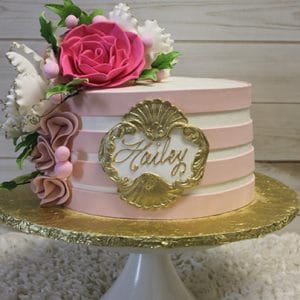 The BR-021 is a round cake with light pink and white stripes, adorned with pink and white fondant flowers, greenery, and a gold ornate frame featuring the name Hailey in elegant script. It sits on a gold board atop a white stand.