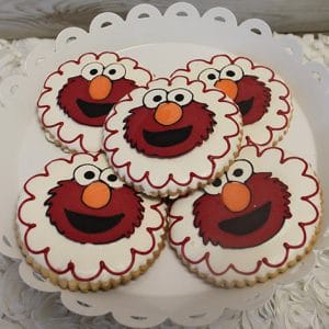 A white plate displays six CE-001d birthday cookies shaped like Elmo's face, decorated with his red fur, big eyes, and orange nose, and outlined with scalloped white icing.