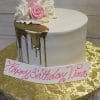 The BR-034 Gold Drip Partial is a white cake with gold drip icing, decorated with pink and white roses on top. It sits on a gold floral-patterned board and features a white plaque reading “Happy Birthday Tina” in pink script.