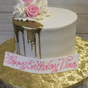 The BR-034 Gold Drip Partial is a white cake with gold drip icing, decorated with pink and white roses on top. It sits on a gold floral-patterned board and features a white plaque reading “Happy Birthday Tina” in pink script.