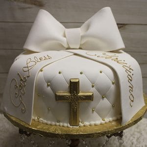The RC-061 cake is a round white cake featuring quilted detailing with small gold accents, a large fondant bow, a gold cross, and gold writing that says "God Bless" and "Christiano.
