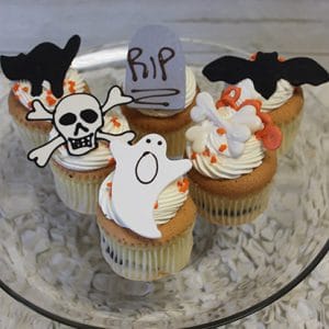 Four CC-051 Display Halloween Gold cupcakes with chocolate filling, each topped with white frosting and Halloween decorations—a black cat, RIP gravestone, bat, skull and crossbones, ghost, and bones—are arranged on a glass plate.