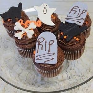 Six CC-050 Display Halloween Chocolate cupcakes with white mousse filling are topped with chocolate frosting, fondant ghosts, bones, tombstones reading "RIP," and orange and black sprinkles for a festive touch on a clear glass plate.