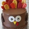THX-030 is a round chocolate cake designed as a turkey, featuring colorful candy feathers on top, large white eyes, a yellow beak, and a red wattle on the front.
