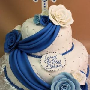 TC-133: Two-tier white cake with blue and white roses, blue drape, decorative cross topper, and “God Bless Ethan” plaque, presented on a silver board.