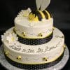 The TC-145 is a two-tier white cake with daisies, small bees, black polka dot ribbon, a fondant bee topper, and "What Will it Bee?" written in cursive on the side.