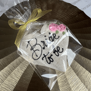 The CE-003h bridal cookie is heart-shaped, decorated with pink roses and "Bride to be" in black icing, wrapped in clear plastic with a gold ribbon, and displayed on a shiny textured gold surface.