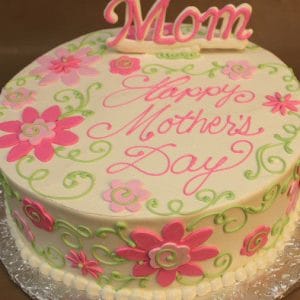 The MD-020 is a round cake with white icing, pink and green floral designs, "Happy Mothers Day" written in pink, and a pink "Mom" topper.