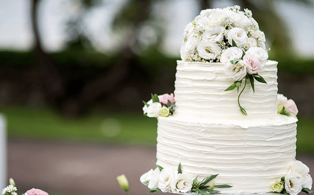 A two-tiered white wedding cake decorated with textured frosting and adorned with white and pale pink flowers, set outdoors with greenery blurred in the background.