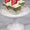 Matcha cake squares topped with cream frosting and fresh strawberries on a white cake stand