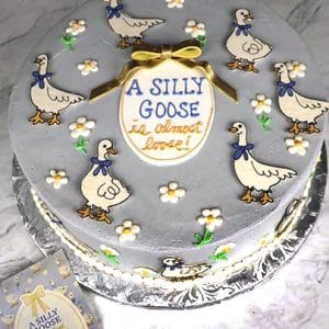 Gray cake decorated with cartoon geese, small flowers, and a message reading “A Silly Goose is almost done”